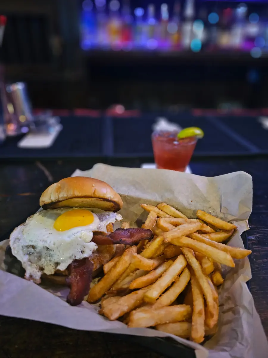 A burger topped with bacon and a fried egg, served in a basket with fries and a cocktail behind it