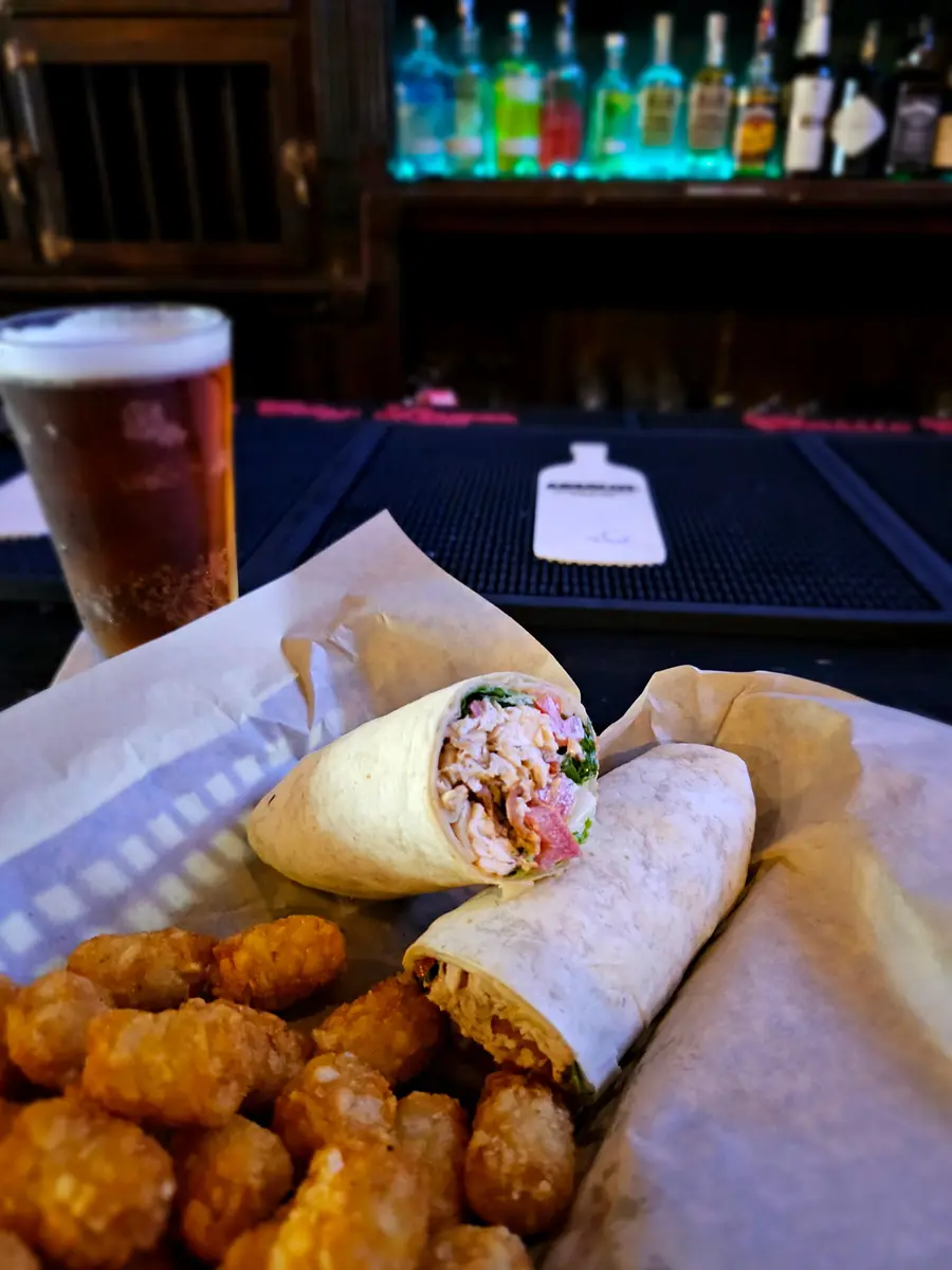 A grilled wrap cut in half with a basket of tots and a pint of amber beer at the bar